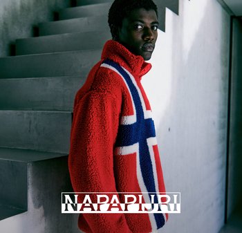 Giovane che indossa una giacca in fleece rossa con un grande design incrociato blu e bianco, in piedi contro una scala di cemento grigia.