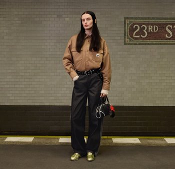 Giovane con capelli lunghi e scuri che indossa una camicia marrone, pantaloni neri larghi, scarpe dorate, tiene una borsa nera e si trova al binario della metropolitana 23rd St.