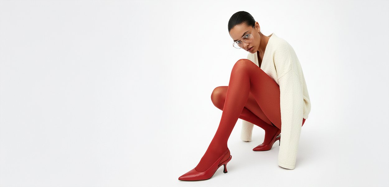 Mujer con gafas que lleva un suéter blanco, medias color óxido y tacones a juego, arrodillada frente a un fondo blanco liso.