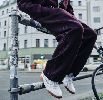 Persona con pantalones morados oscuros y zapatillas blancas Reebok sentada en una barandilla de metal, con una bicicleta cerca, en una calle de la ciudad.