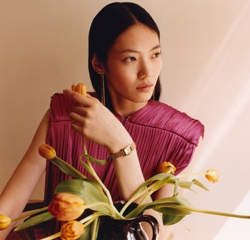 Top fucsia plissé con spalle larghe, orologio d'oro al polso, lunghi orecchini d'oro e tulipani gialli in un vaso nero. Luce sfumata sullo sfondo.