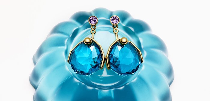 Boucles d’oreilles en or avec de grandes pierres précieuses bleues facettées et de petites pierres violettes, présentées sur un plat bleu brillant à festons.