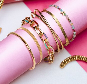 Braccialetti tono oro di design vario, inclusi una catena e fasce lisce, insieme a un braccialetto di perline con una lettera, disposti su carta rosa.