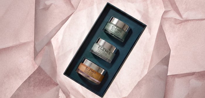 Trois pots de soins Elemis Pro-Collagen — crème de nuit, crème marine SPF 30, baume nettoyant — disposés dans une boîte noire sur du papier de soie rose.