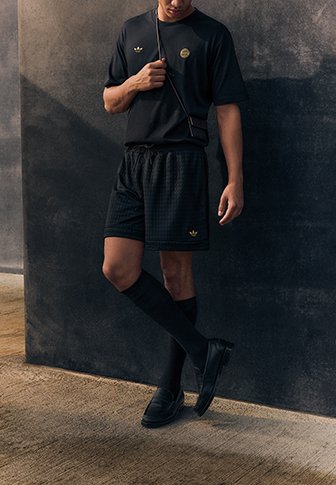 Man die een zwart sportoutfit met een gouden logo draagt, met één been gekruist staat en leunt tegen een donkere, gestructureerde muur.