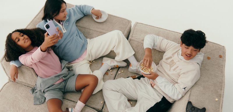 Trois enfants se prélassent sur un canapé gris. Ils portent des tenues décontractées : un pull en polaire bleu clair, des hauts rose et gris, et un pantalon blanc. Des collations sont présentes.