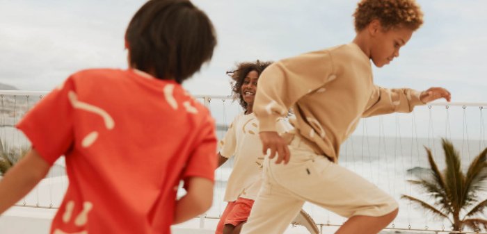 Two girls stand in a field wearing white skirts with colorful floral embroidery; one wears an orange top, the other a white dress with flutter sleeves.