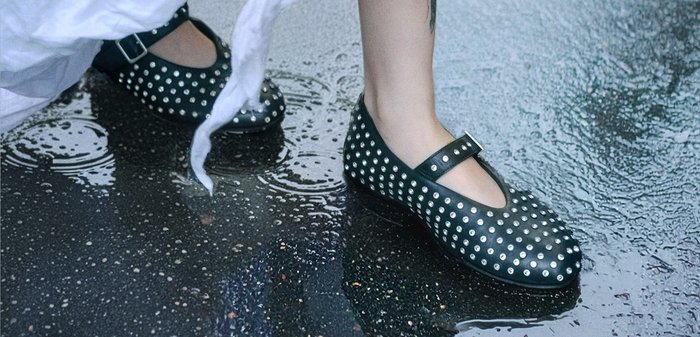 Zwarte leren Mary Jane-schoenen met zilveren studs, voorzien van een gespband en een ronde neus, geplaatst op een natte ondergrond met reflecties.