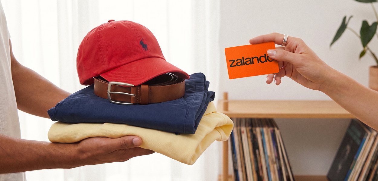Une personne tient des vêtements pliés et des accessoires tandis qu'une autre personne propose une carte Zalando orange pour le paiement ou comme cadeau.