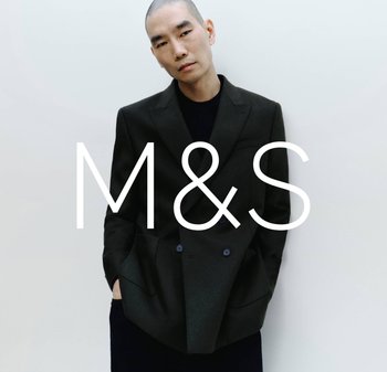 Mężczyzna w ciemnej marynarce dwurzędowej i czarnej koszuli stoi na jasnym tle z logo M&S na zdjęciu.