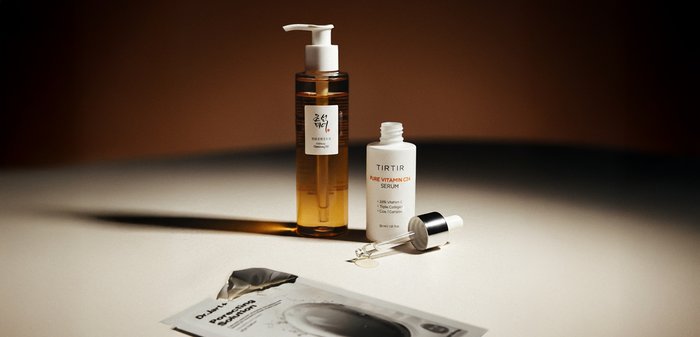 Huile démaquillante ambrée avec pompe, flacon blanc de sérum à la vitamine C avec compte-gouttes, et sachet masque tissu argenté pour soins de la peau sur une surface claire.