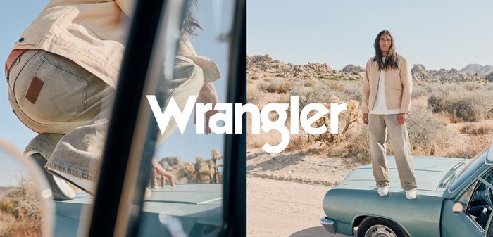 Man in beige jas en broek staat op de motorkap van een vintage blauwe auto in een woestijnlandschap met rotsachtige heuvels en droge begroeiing.