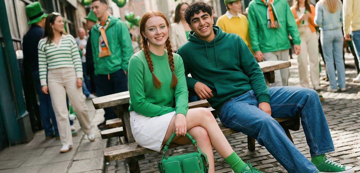 Two young people dressed in green sit on a wooden bench, smiling, with a festive crowd wearing green behind them in a street setting.