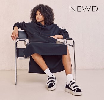 Black hooded dress with a relaxed fit, worn with patterned socks and chunky black-and-white sneakers, seated in a modern chair.