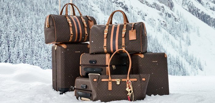 Cuatro bolsas de viaje marrones con estampado y detalles en cuero beige, con herrajes dorados, apiladas sobre la nieve frente a un fondo de bosque.