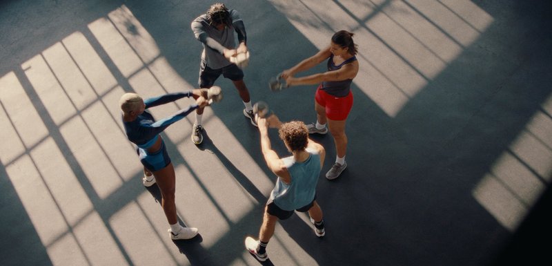 Τέσσερα άτομα γυμνάζονται με αλτήρες kettlebell σε έναν ηλιόλουστο γυμναστήριο, στέκονται σε κύκλο και κουνάνε τα βάρη προς τα εμπρός ταυτόχρονα.