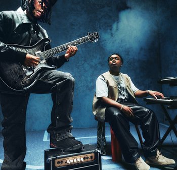 Un homme joue de la guitare électrique avec le pied posé sur un ampli, tandis qu'un autre homme est assis au clavier dans une pièce enfumée éclairée en bleu.