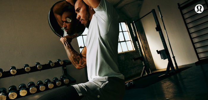 Atletische man die een gewichtsschijf optilt, gekleed in een aansluitend wit shirt en grijze korte broek, met zichtbare gespierde armen. Fitnessapparatuur op de achtergrond.
