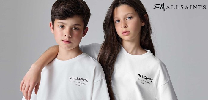 Chlapec a dívka stojí vedle sebe, oba mají na sobě bílé trička AllSaints, chlapcova ruka spočívá na dívčině rameni, šedé pozadí.