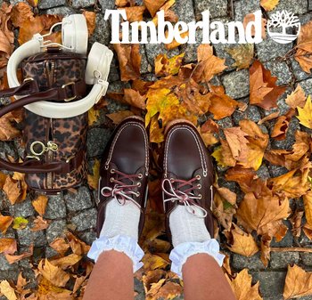 Braune Timberland-Lederschuhe mit weißen Schnürsenkeln, getragen mit strukturierten weißen Socken und gerafften Bündchen, auf einem Bett aus Herbstblättern und Steinen.