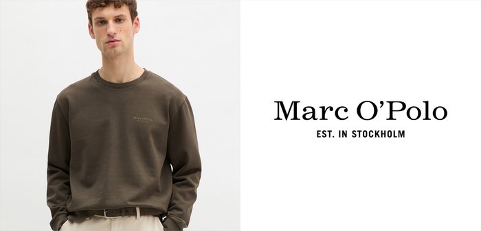 Młody mężczyzna w ciemnooliwkowej bluzie i beżowych spodniach stojący na tle białej ściany z napisem „Marc O’Polo EST. W Sztokholmie”.