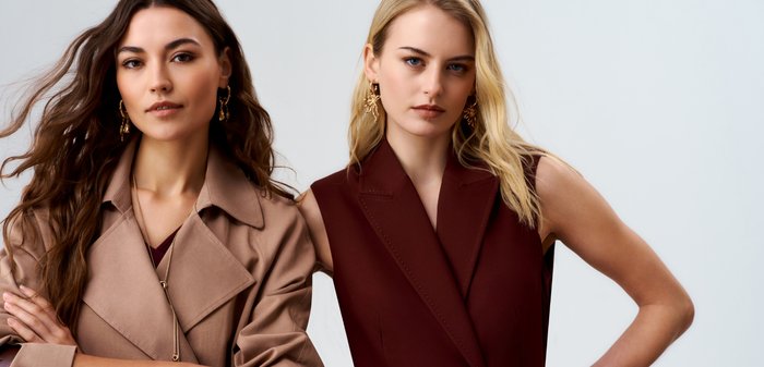 Twee vrouwen dragen mouwloze, met een ceintuur voorziene jassen, één beige en één donkerrood, met gouden oorbellen, staan zelfverzekerd tegen een effen lichte achtergrond.