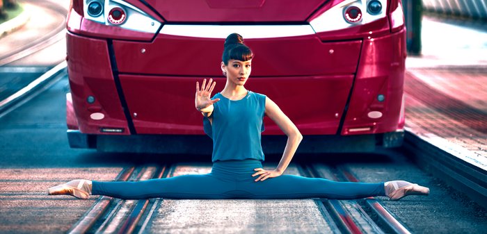 Een rood openbaarvervoerbusje staat tegenover een danseres in een blauw kostuum, die een spreidsprong uitvoert op een spoor. Op de achtergrond zijn vage stedelijke elementen te zien.