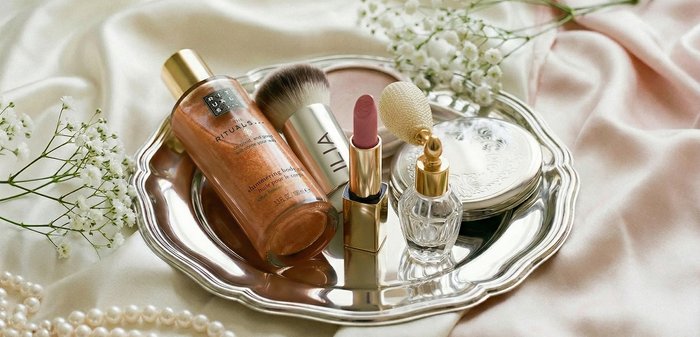 Plateau en argent contenant une bouteille d'huile corporelle scintillante, un pinceau à maquillage, un rouge à lèvres rose, un atomiseur de parfum vintage et un miroir compact sur tissu satin.