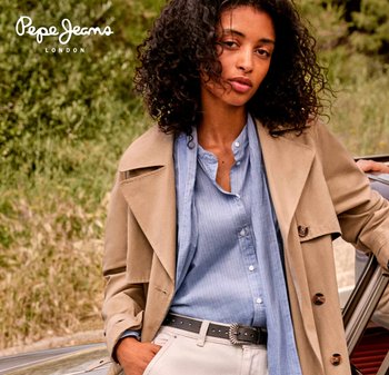 Trench beige sobre una camisa de botones azul clara. La camisa tiene rayas verticales. Cinturón negro con hebilla plateada. El fondo es de vegetación.
