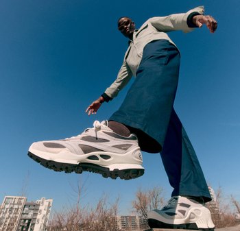 Weisse und graue Sportschuhe mit einer strukturierten Sohle, die schwarze Akzente und Schnürsenkel aufweisen. Kombiniert mit blauen weit geschnittenen Hosen und einer grünen Jacke.