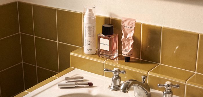 Flacon de parfum, spray de soin de la peau et tube de crème froissé sur un lavabo de salle de bain avec un mur carrelé jaune moutarde et des poignées de robinet en argent.