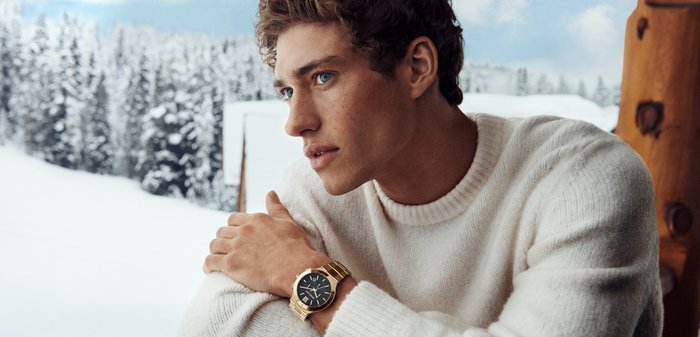 Guldarmbandsur med svart urtavla och romerska siffror. Bärs av en person i en krämfärgad stickad tröja, med snöigt landskap i bakgrunden.