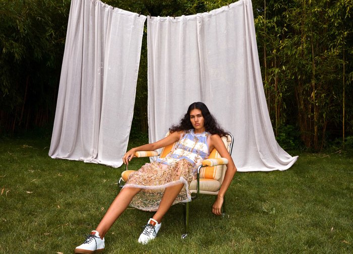 Jonge vrouw met lang donker haar zittend buiten in een gestreepte fauteuil, gekleed in een doorschijnende gouden borduursel jurk en witte schoenen op het gras.