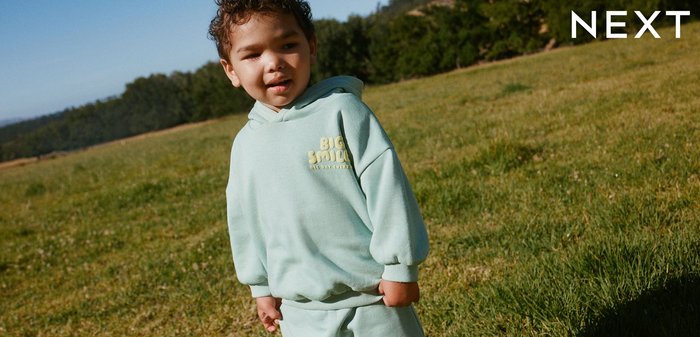 Malé dieťa oblečené v svetlomodrej mikine a šortkách stojace na trávnatej lúke s lesom v pozadí.