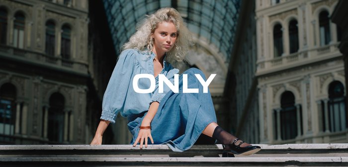 Tânără cu păr blond creț, purtând un costum din denim albastru, stă încrezătoare într-un arcade urban cu tavan de sticlă, cu textul "ONLY" suprapus.