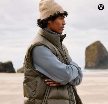 Olijfgroene gewatteerde bodywarmer over een lichtblauwe trui, met een beige gebreide muts. Op de achtergrond is een strand en rotsformaties te zien.