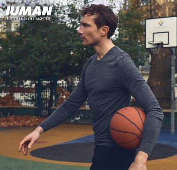 Uomo atletico con una maglia slim fit a maniche lunghe di colore grigio scuro, che tiene un pallone da basket sotto il braccio, in piedi su un campo all'aperto colorato.