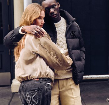 Två unga vuxna, en i en beige jacka och mörka jeans, den andra i en svart puffjacka och en krämfärgad tröja, kramas framför en mörk bakgrund.