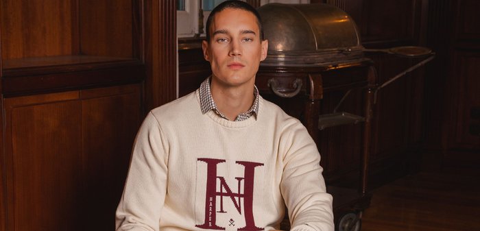 Kremowy sweter z dzianiny z bordowym logo, w zestawieniu z koszulą w kratę, niebieskimi dżinsami i zegarkiem. Otoczony stosami książek na drewnianej podłodze.