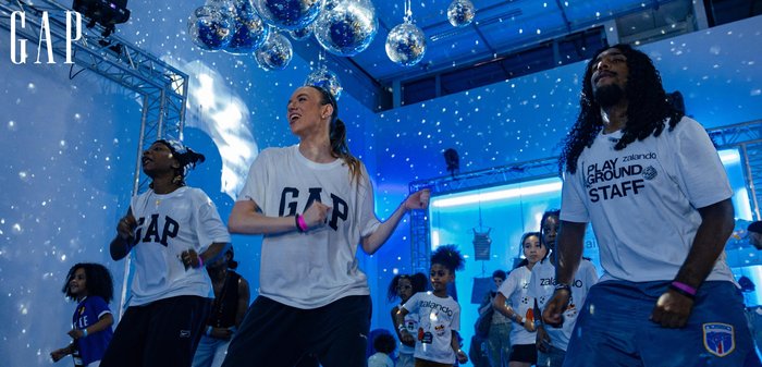 Groupe de personnes diverses dansant à l'intérieur sous des boules disco suspendues avec un éclairage bleu, portant des t-shirts de marques GAP et Zalando.