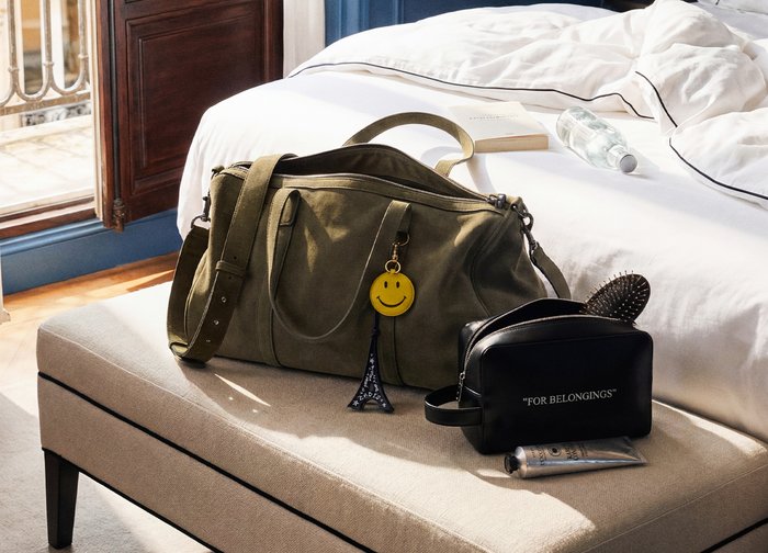 Πράσινη τσάντα ταξιδιού με μπρελόκ με χαμογελαστό πρόσωπο και Πύργο του Άιφελ, μαύρη τσάντα για είδη προσωπικής υγιεινής, βούρτσα μαλλιών και λοσιόν πάνω σε πάγκο υπνοδωματίου κοντά στο κρεβάτι.