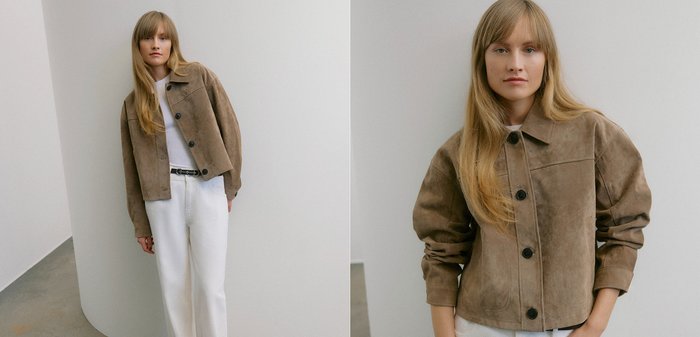 Jonge vrouw met lang blond haar, gekleed in een bruine jas met knopen, een wit topje, witte broek en een zwarte riem, staand tegen een gebogen witte muur.