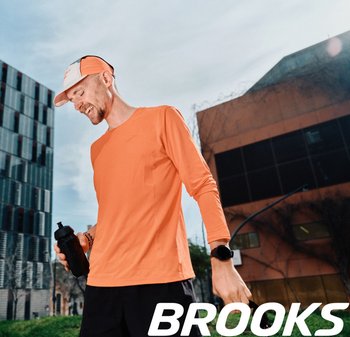 Langermige oranje sportshirt gemaakt van ademend materiaal, gecombineerd met zwarte shorts en een zwarte waterfles. Buitenscene in de stad.