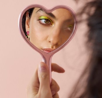 Donna che tiene uno specchio rosa a forma di cuore che riflette il suo volto con ombretto dorato e orecchini floreali su uno sfondo rosa pallido.