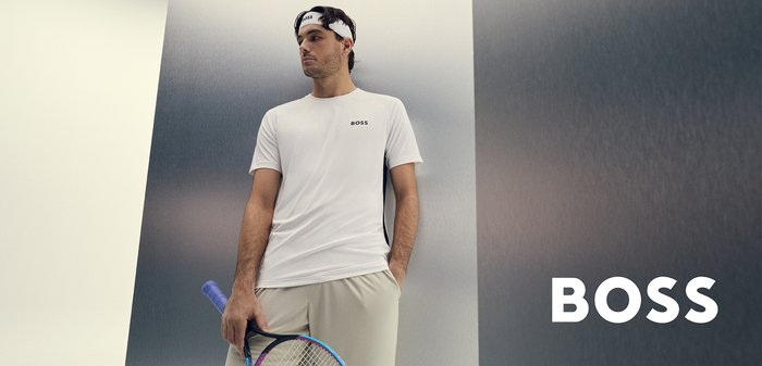 Hombre con ropa deportiva blanca de BOSS y banda para la cabeza sostiene una raqueta de tenis, posando sobre un fondo degradado gris y blanco con el logo de BOSS.