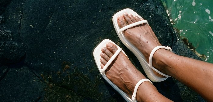Pieds à peau mouillée portant des sandales blanches à brides, reposant sur des rochers sombres à côté d'eau verte.