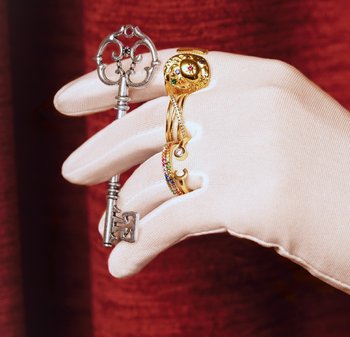 Zilveren sleutel met versierde sluier, gecombineerd met een gouden ring met kleurrijke stenen en een spiraalontwerp, vastgehouden door een handschoen tegen een rode achtergrond.