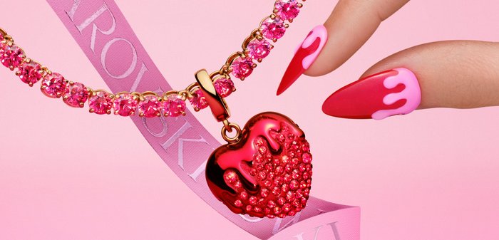 Hand met glanzende roze en rode nagels die een hartvormige hanger van roze kristal vasthoudt aan een bijpassende roze kristallen ketting, over een roze Swarovski-lint.