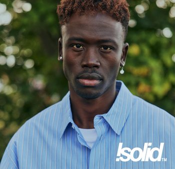 Junger schwarzer Mann mit kurzen lockigen Haaren und Ohrringen, trägt ein blau gestreiftes Hemd, steht im Freien vor unscharfem grünem Laub im Hintergrund.