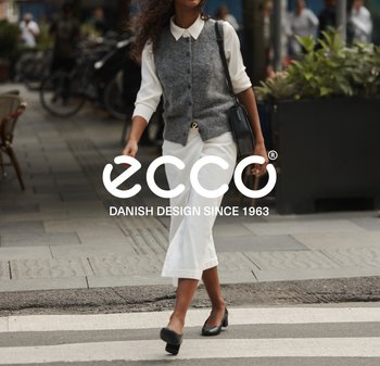 Woman walking across a street crosswalk wearing black heels, white wide-leg pants, white shirt, grey vest, and carrying a black handbag.